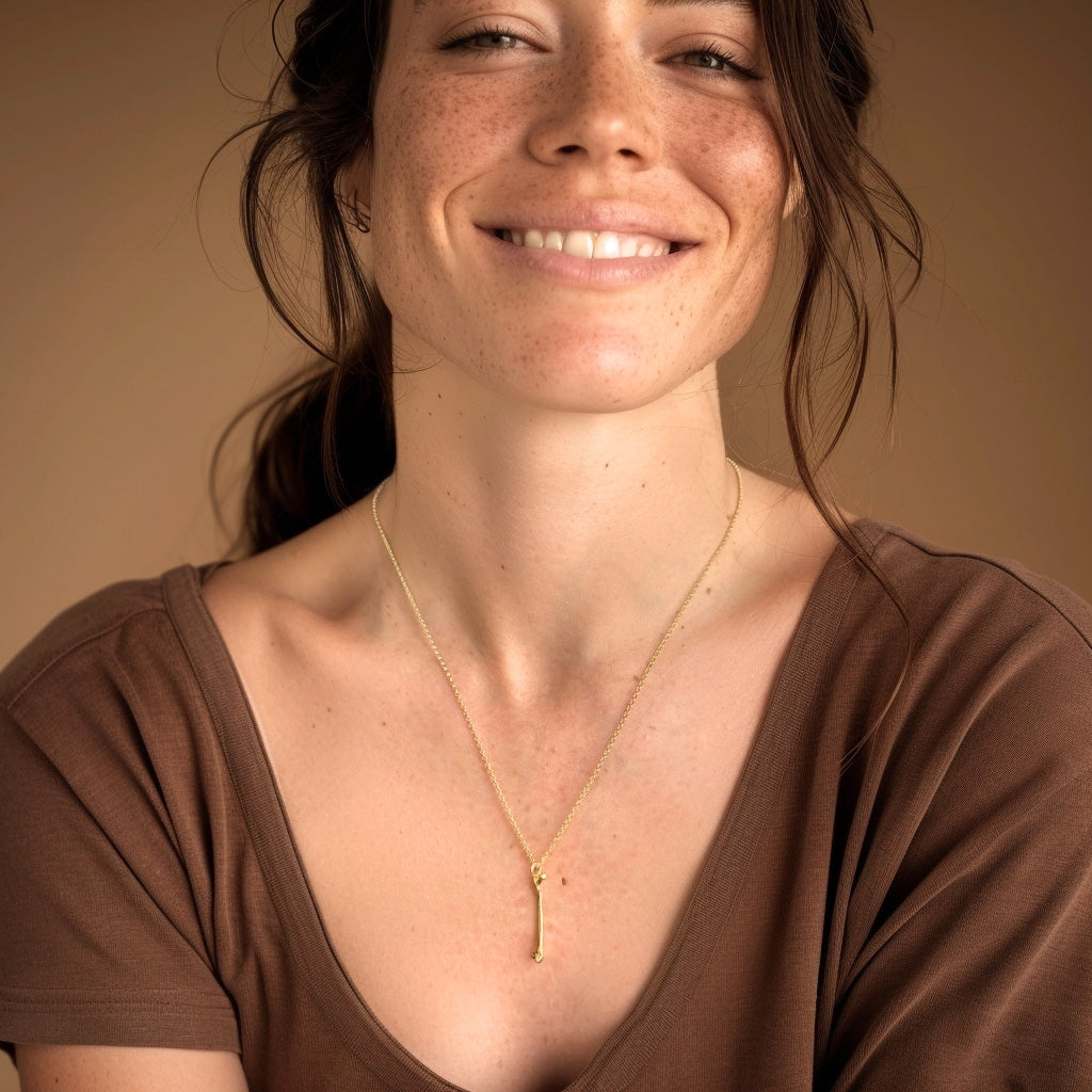 A smiling woman wearing the gold femur necklace over a brown top, the pendant hanging at chest level and catching the light. A smiling woman wearing the gold femur necklace over a brown top, the pendant hanging at chest level and catching the light.