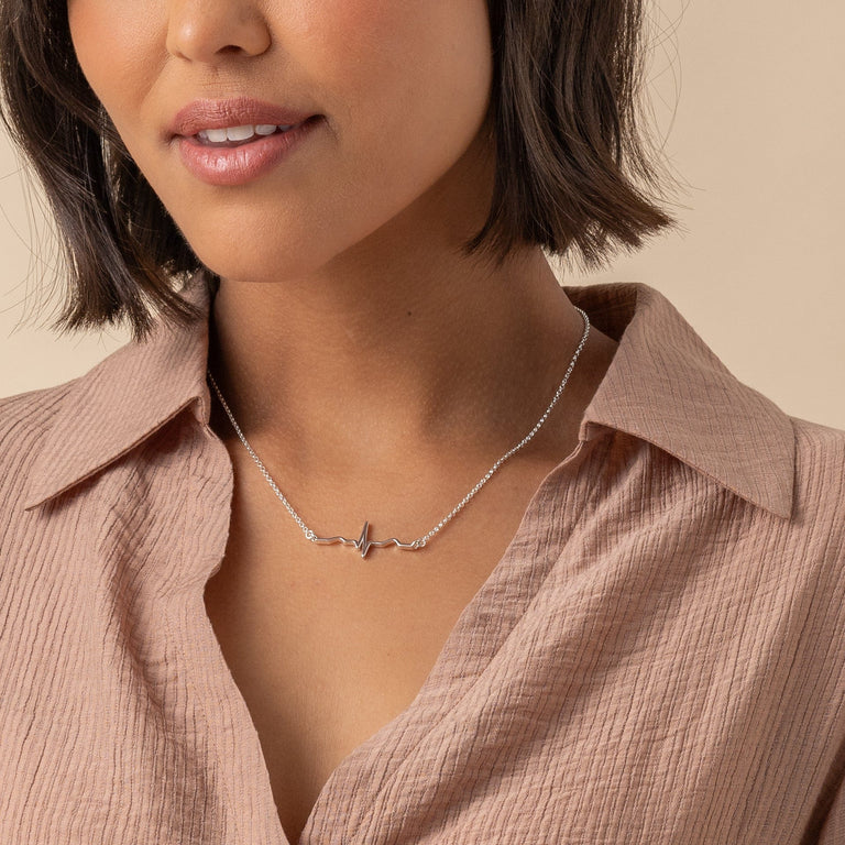 A model wearing a sterling silver heartbeat necklace, showcasing a minimalist design with a heartbeat line pendant against a soft pink blouse. The delicate chain complements the modern and elegant look of the jewelry. A model wearing a sterling silver heartbeat necklace, showcasing a minimalist design with a heartbeat line pendant against a soft pink blouse. The delicate chain complements the modern and elegant look of the jewelry.