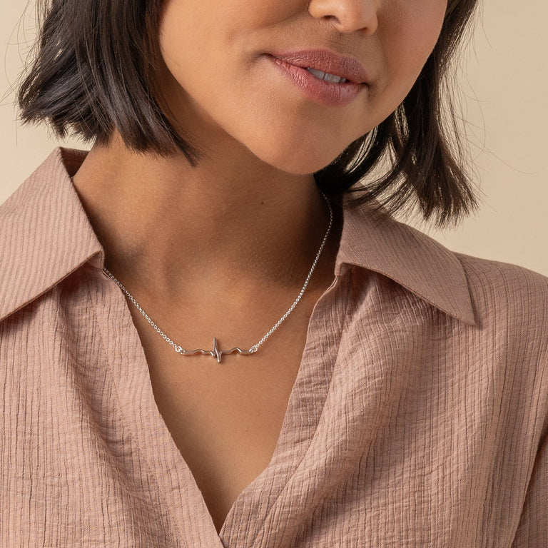 A sterling silver heartbeat necklace worn by a model, paired with a beige blouse. The heartbeat line pendant elegantly adorns the neckline, adding a touch of delicate and contemporary style to the outfit. A sterling silver heartbeat necklace worn by a model, paired with a beige blouse. The heartbeat line pendant elegantly adorns the neckline, adding a touch of delicate and contemporary style to the outfit.