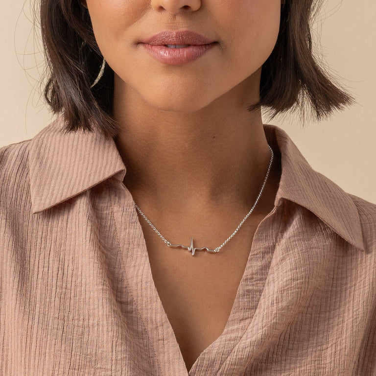 Close-up of a model wearing a sterling silver heartbeat necklace. The necklace features a heartbeat line pendant, elegantly resting on the collarbone, paired with a light, textured blouse for a chic and sophisticated style. Close-up of a model wearing a sterling silver heartbeat necklace. The necklace features a heartbeat line pendant, elegantly resting on the collarbone, paired with a light, textured blouse for a chic and sophisticated style.