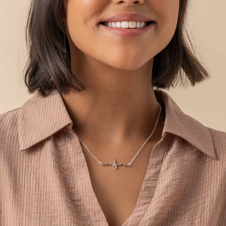 Sterling silver heartbeat necklace displayed on a model. The necklace, featuring a heartbeat line design, stands out against a soft pink blouse, highlighting its sleek and stylish appeal for a modern, science-inspired look. Sterling silver heartbeat necklace displayed on a model. The necklace, featuring a heartbeat line design, stands out against a soft pink blouse, highlighting its sleek and stylish appeal for a modern, science-inspired look.