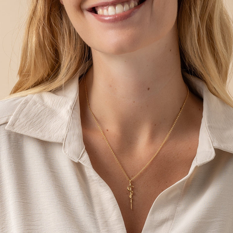 Woman wearing the gold vermeil Rod of Asclepius necklace, highlighting its sleek and meaningful style for healthcare professionals. Woman wearing the gold vermeil Rod of Asclepius necklace, highlighting its sleek and meaningful style for healthcare professionals.