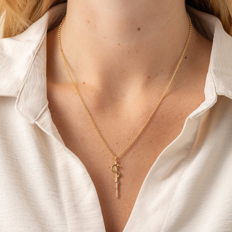 Side view of a model wearing the gold vermeil Rod of Asclepius necklace, a symbol of healthcare and renewal. Side view of a model wearing the gold vermeil Rod of Asclepius necklace, a symbol of healthcare and renewal.