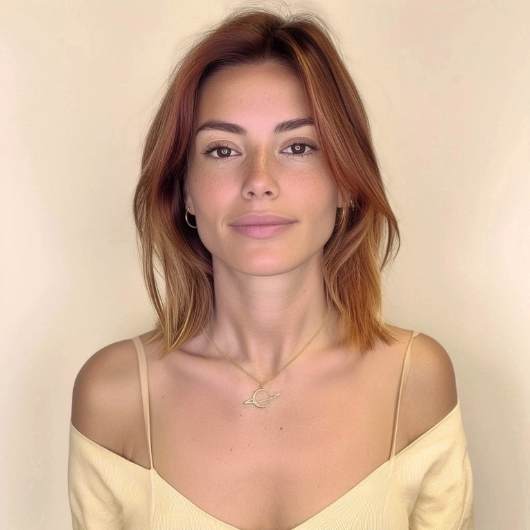 Woman with shoulder-length auburn hair wearing a gold Saturn necklace, shown from the waist up, against a soft beige background. Woman with shoulder-length auburn hair wearing a gold Saturn necklace, shown from the waist up, against a soft beige background.