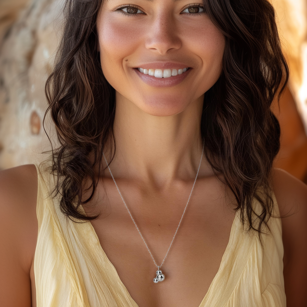 A smiling woman with wavy dark hair wearing a sterling silver H₂O molecule necklace against a warm, natural backdrop, showcasing the elegant and scientific design. A smiling woman with wavy dark hair wearing a sterling silver H₂O molecule necklace against a warm, natural backdrop, showcasing the elegant and scientific design.