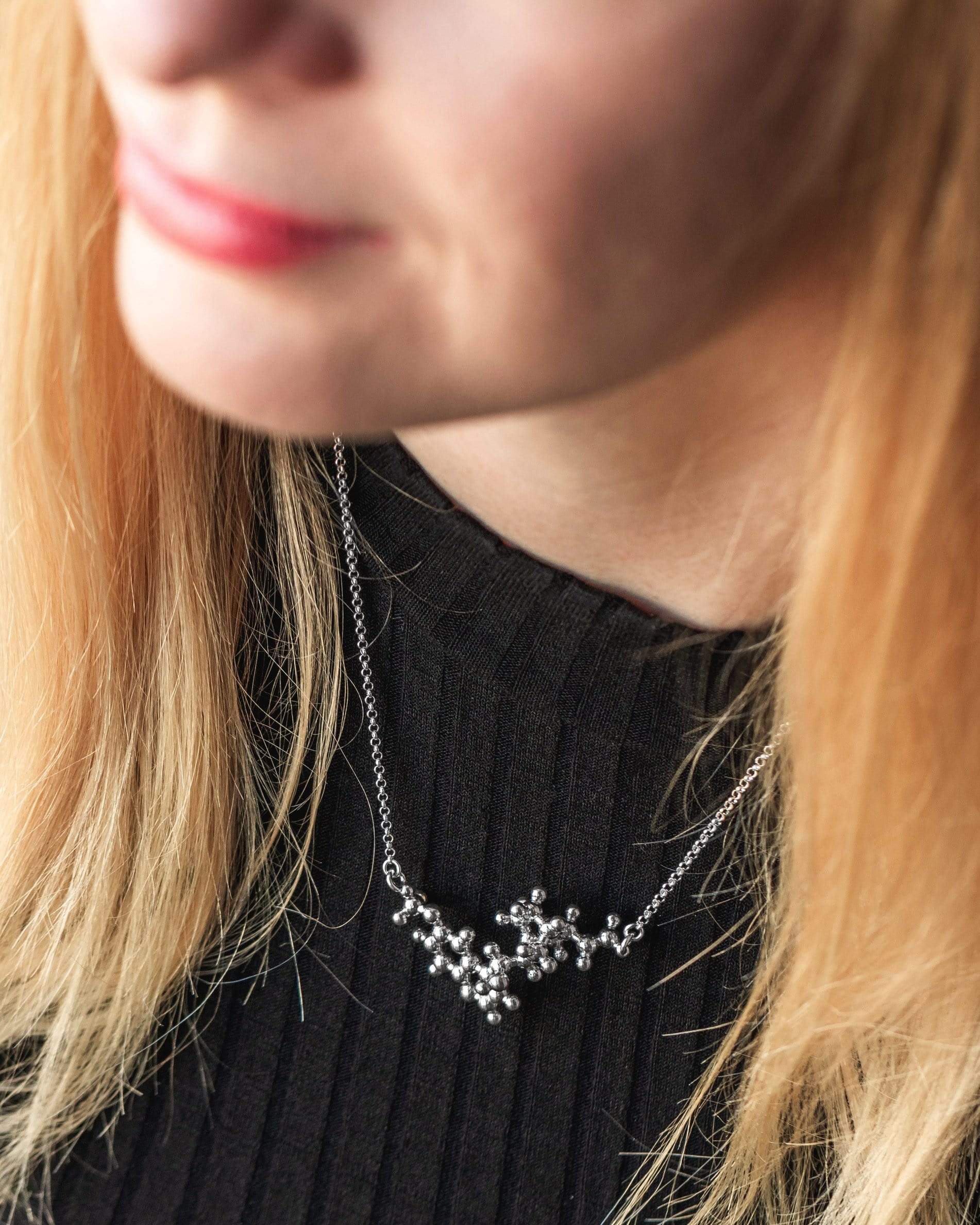 Model wearing the quinine necklace in sterling silver, showcasing the detailed molecular structure against a black outfit. Model wearing the quinine necklace in sterling silver, showcasing the detailed molecular structure against a black outfit.