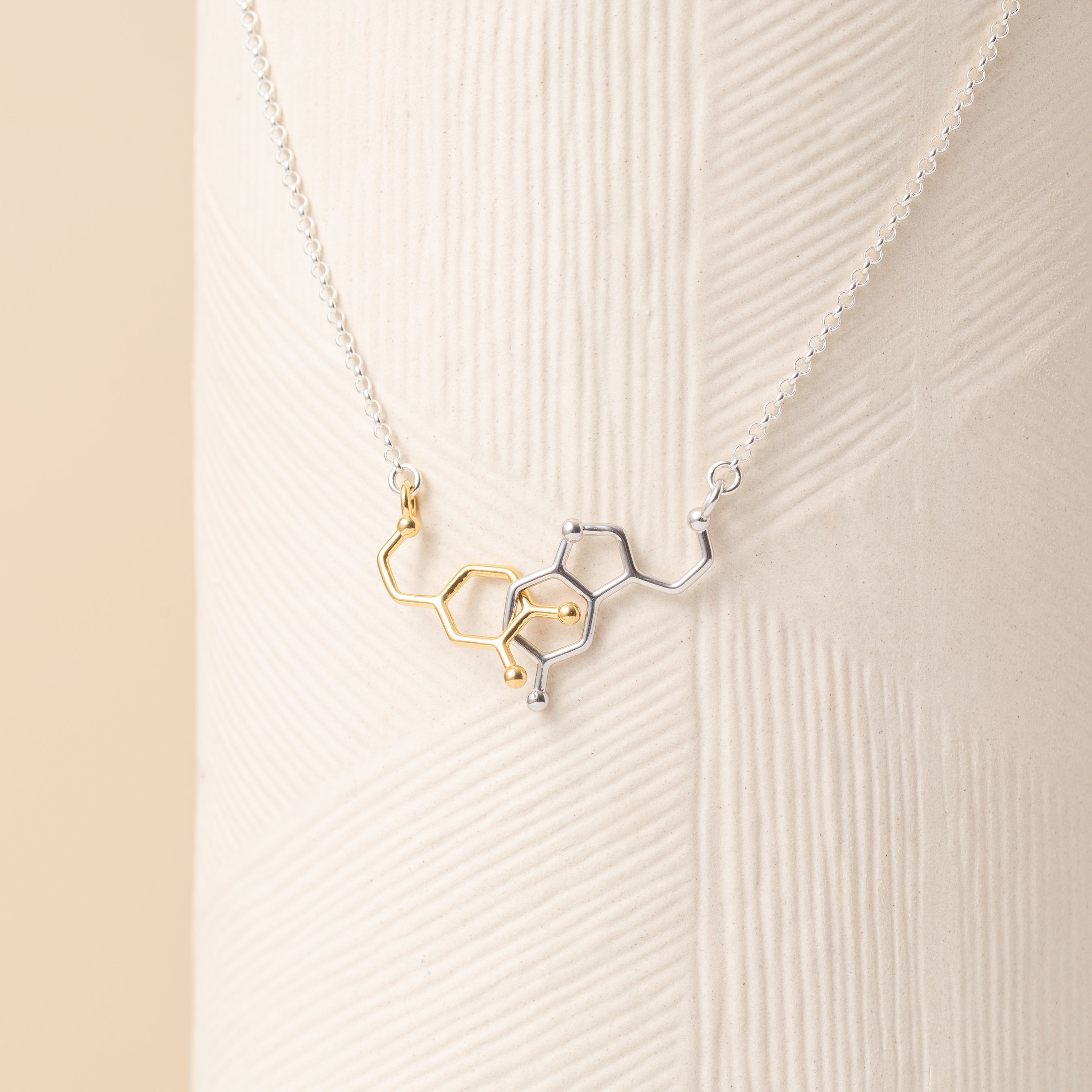 Close-up of interlocked dopamine and serotonin molecule necklace in mixed metals—gold vermeil and polished sterling silver—displayed on a textured neutral background