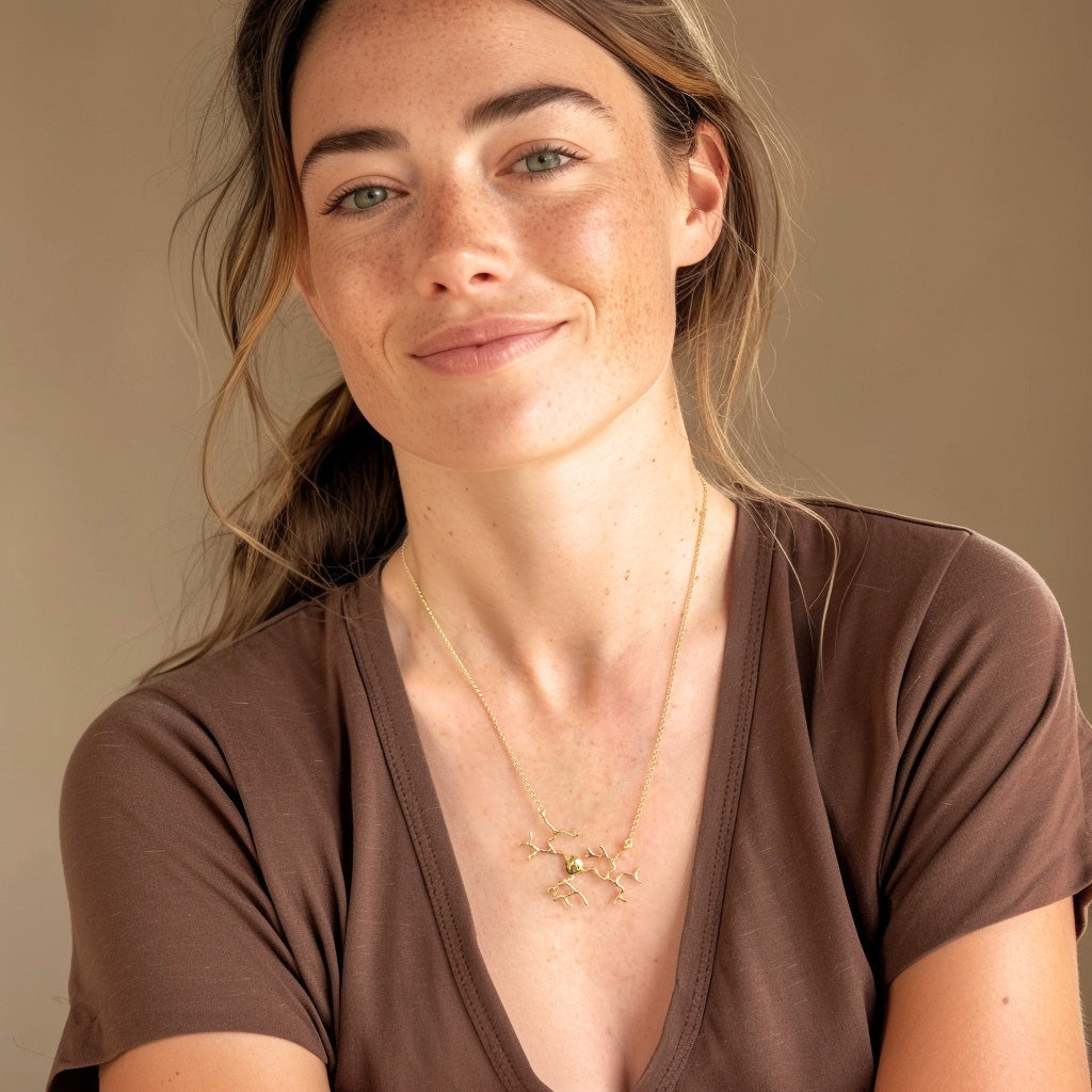 Woman with light skin and long brown hair wearing a gold vermeil neuron necklace, smiling softly while dressed in a brown V-neck top.