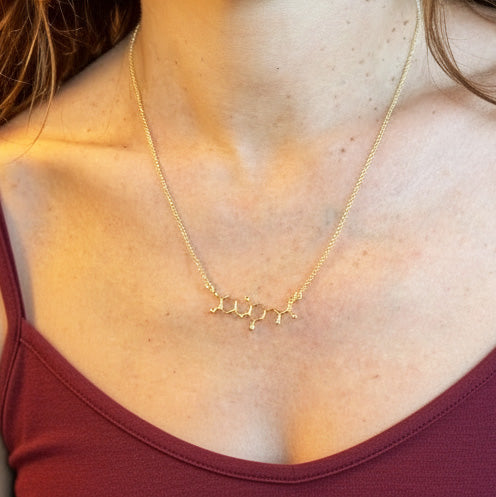 Close-up of a gold vermeil T3 thyroid hormone necklace resting just below the collarbones of a person wearing a burgundy top, highlighting the pendant’s detailed molecular shape.