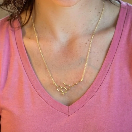 Close-up of a person wearing a gold vermeil THC molecule necklace, resting at the collarbone above a pink V-neck shirt.