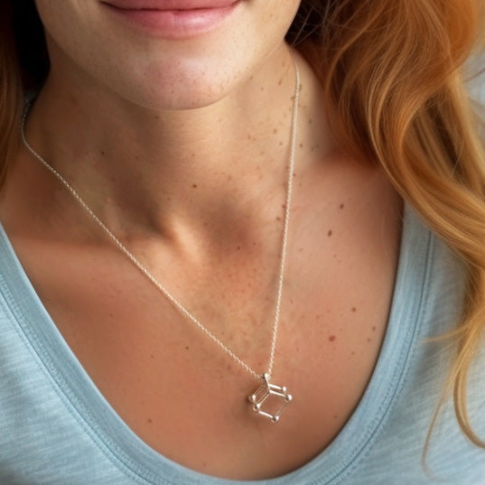 Close-up view of a woman’s neck and upper chest wearing the arsenic trioxide molecular structure necklace on a fine silver chain, paired with a light blue shirt.