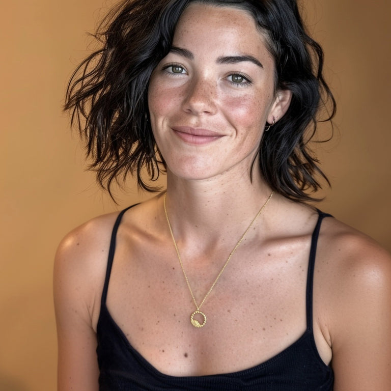 Smiling woman wearing the gold vermeil blastocyst necklace with a circular cell-inspired pendant, styled with a black spaghetti-strap top and warm background.