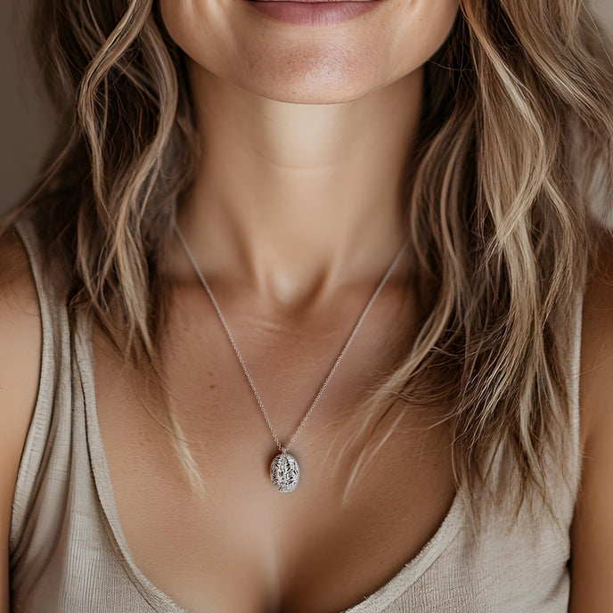 Close-up of a woman wearing a silver brain-inspired pendant necklace on a delicate chain. The necklace features an intricate design resembling a human brain. She is dressed in a beige tank top, and her wavy, light brown hair frames her neck. The background is softly blurred, creating a warm and elegant aesthetic.