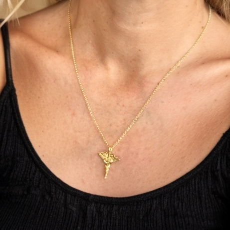 Woman wearing a gold vermeil caduceus necklace on a fine chain, shown against a black top with a warm beige background.