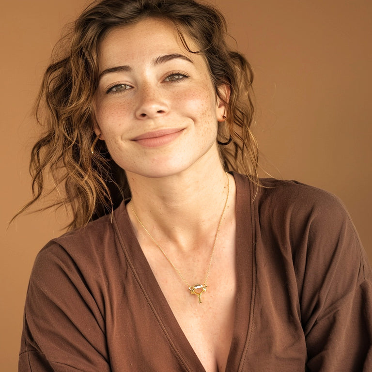 Smiling woman with curly hair wearing the cervical vertebra necklace in gold vermeil, styled with a brown V-neck top against a warm beige background.