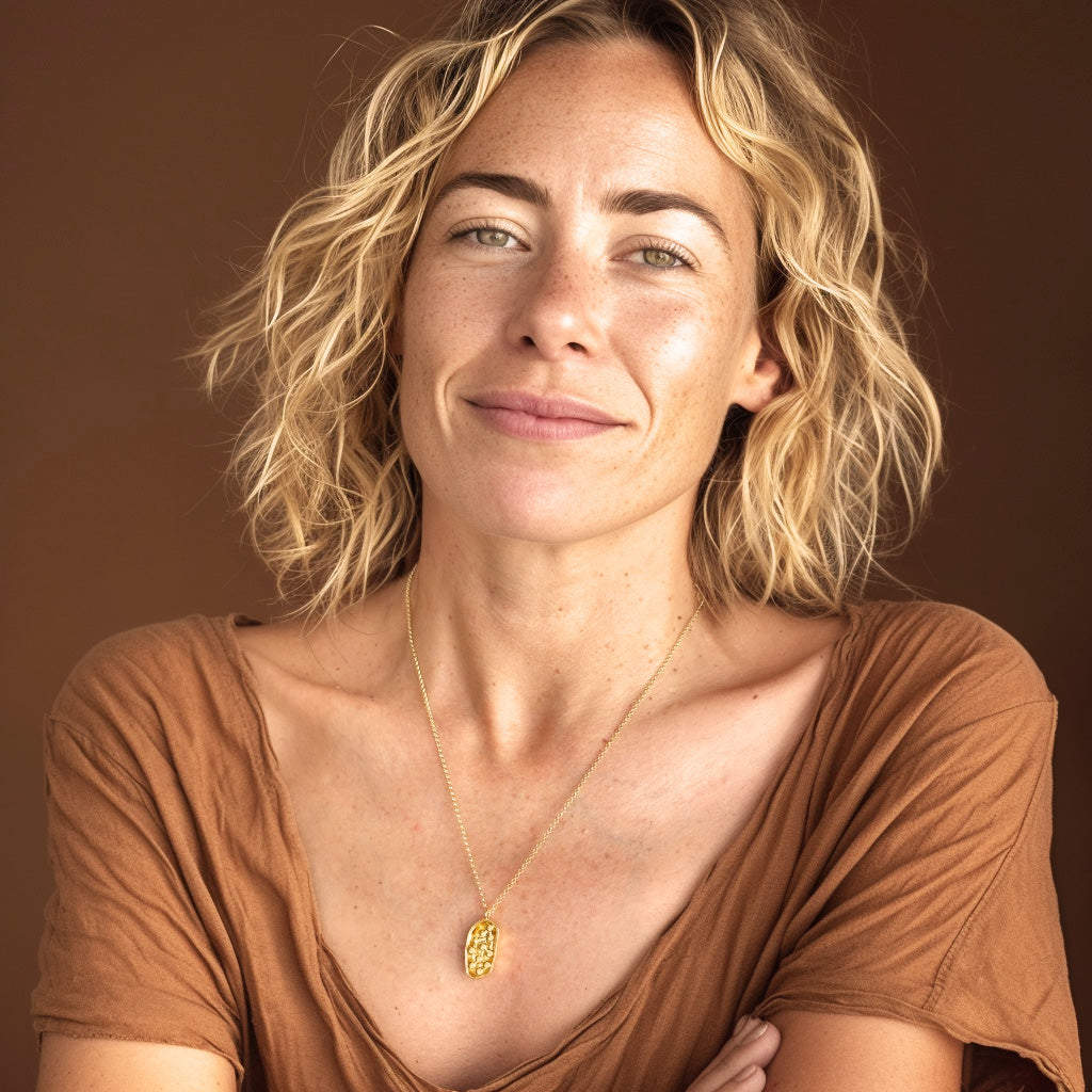 Smiling woman with wavy blonde hair wearing a gold vermeil chloroplast necklace, styled with a warm brown top against a soft background.