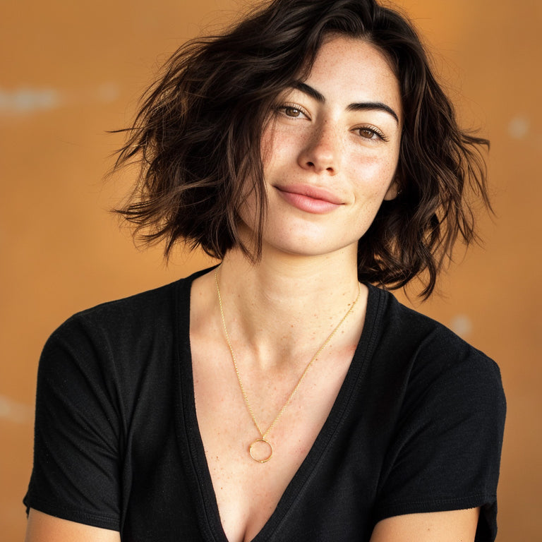Smiling woman with short wavy brown hair wearing a black V-neck top and the Cloning Vector Necklace in gold vermeil against a warm orange background.