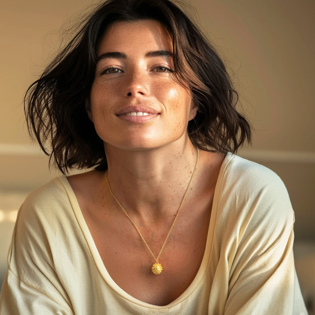 Woman with dark wavy hair wearing a gold vermeil coronavirus necklace, smiling softly while seated indoors in warm lighting, dressed in a cream-colored top.