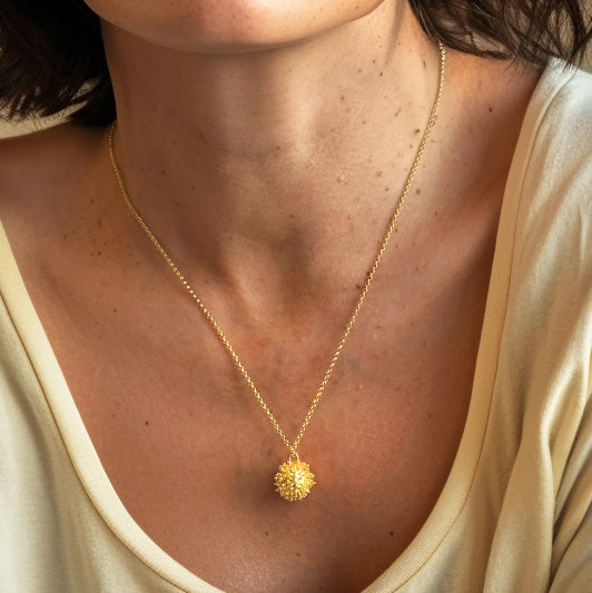 Close-up view of a woman’s neck and chest wearing a gold vermeil coronavirus necklace, with the intricate pendant resting just below the collarbone, paired with a cream-colored shirt.