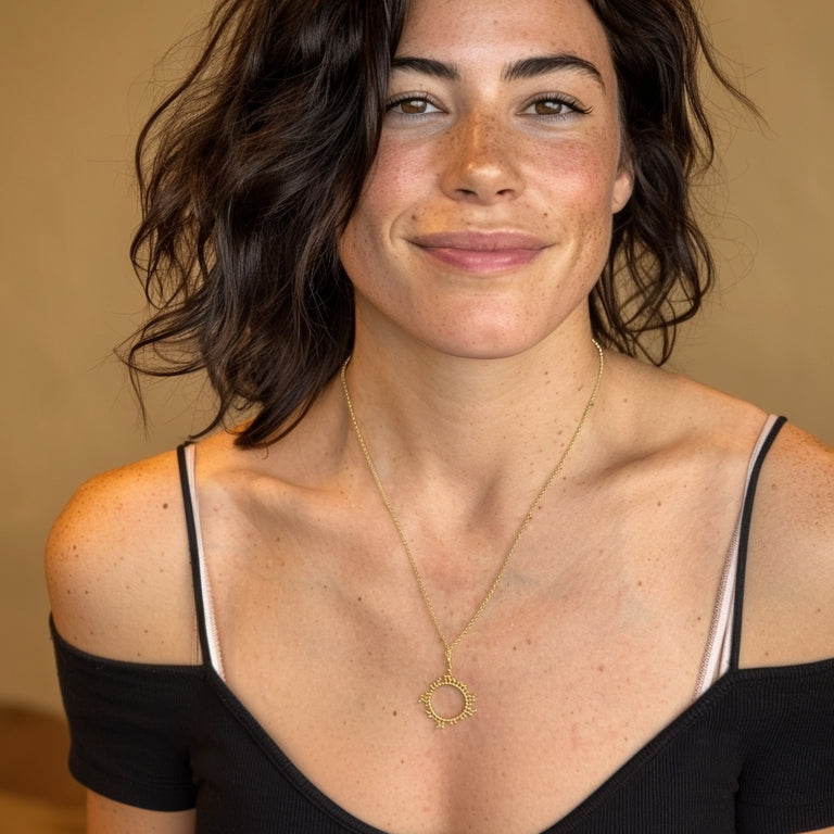 A smiling woman with shoulder-length dark hair wearing the cyclosporine necklace in gold vermeil, styled with an off-shoulder black dress, illustrating how the jewelry looks when worn.