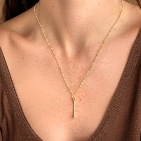 A close-up of the gold femur necklace worn on the neck, the anatomical pendant resting elegantly on the chest against a brown shirt.