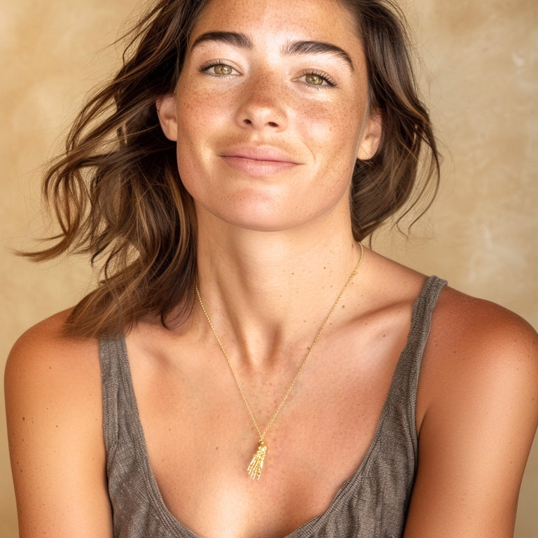Smiling woman with tanned skin and brown hair wearing the gold vermeil foot anatomy necklace, styled with a casual sleeveless top.