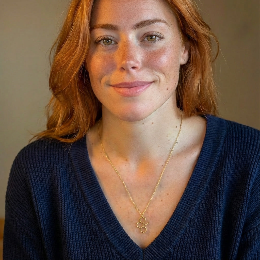Woman wearing a gold vermeil ketamine molecule necklace, shown from the shoulders up, smiling slightly in a blue V-neck sweater