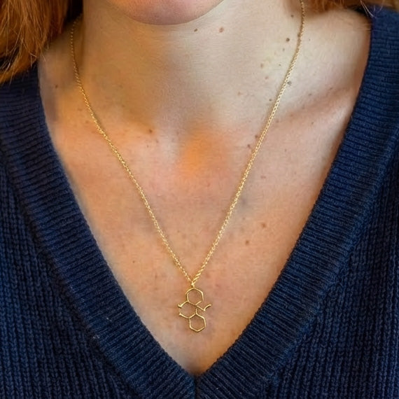 Close-up of the gold vermeil ketamine molecule necklace resting on the chest of a woman wearing a blue V-neck sweater