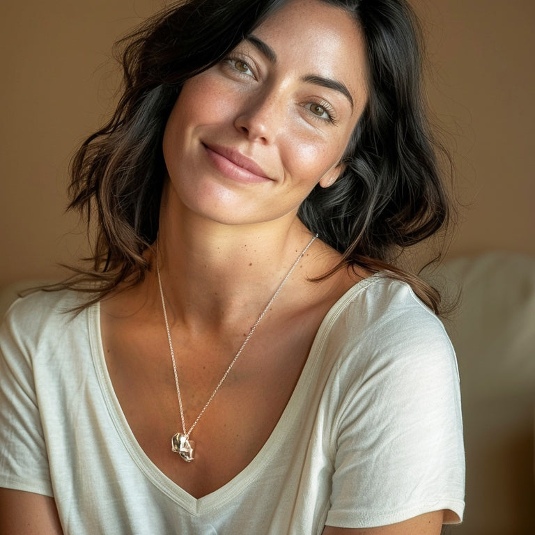 A woman with wavy dark hair wearing the macaque skull necklace and a light-colored V-neck shirt. She smiles gently while seated, showcasing the necklace in a natural, relaxed setting.