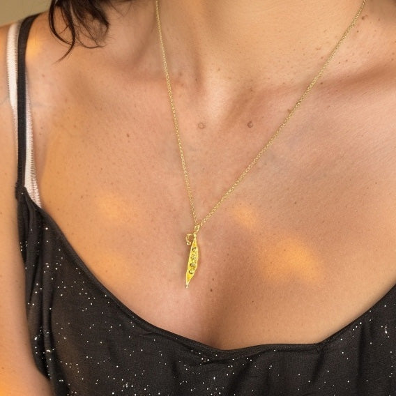 Woman wearing a gold pea pod necklace on a delicate chain, photographed close-up against her skin in warm light.