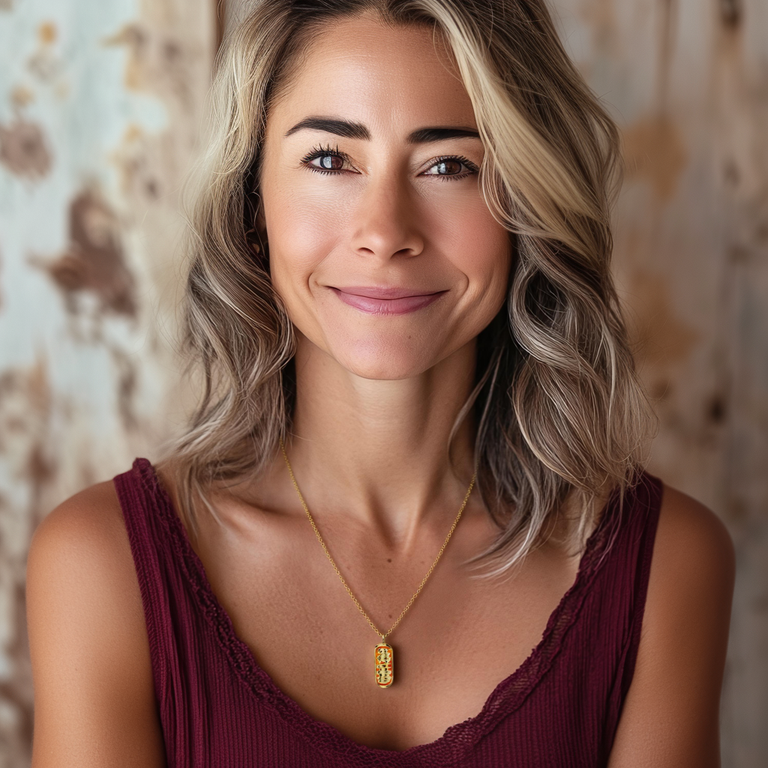 A woman with wavy blonde hair and a warm smile wears the gold vermeil mitochondrion necklace. The pendant hangs elegantly against her skin, complementing her deep burgundy top. A soft-focus background adds to the natural and sophisticated look.