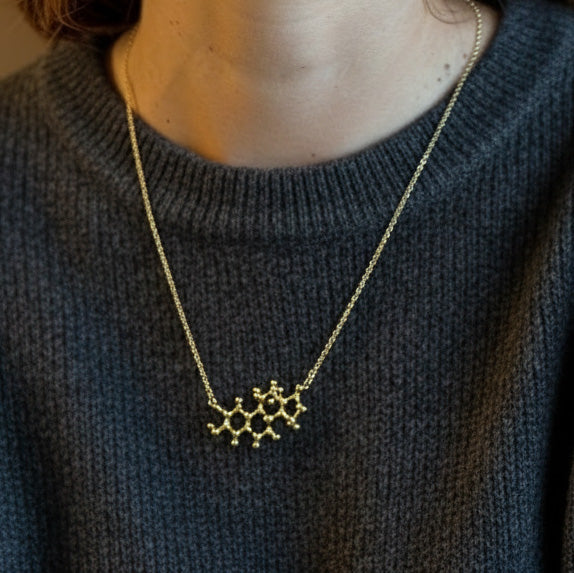 Gold vermeil estrogen molecule necklace worn on the collarbone, shown in a close-up of the neck and upper chest over a dark grey sweater.