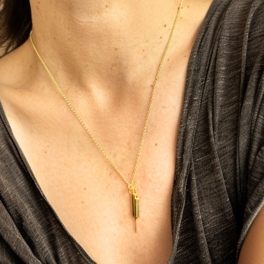 Gold vermeil pipette necklace worn around the neck, close-up view highlighting the pendant’s shiny finish against a grey V-neck shirt.