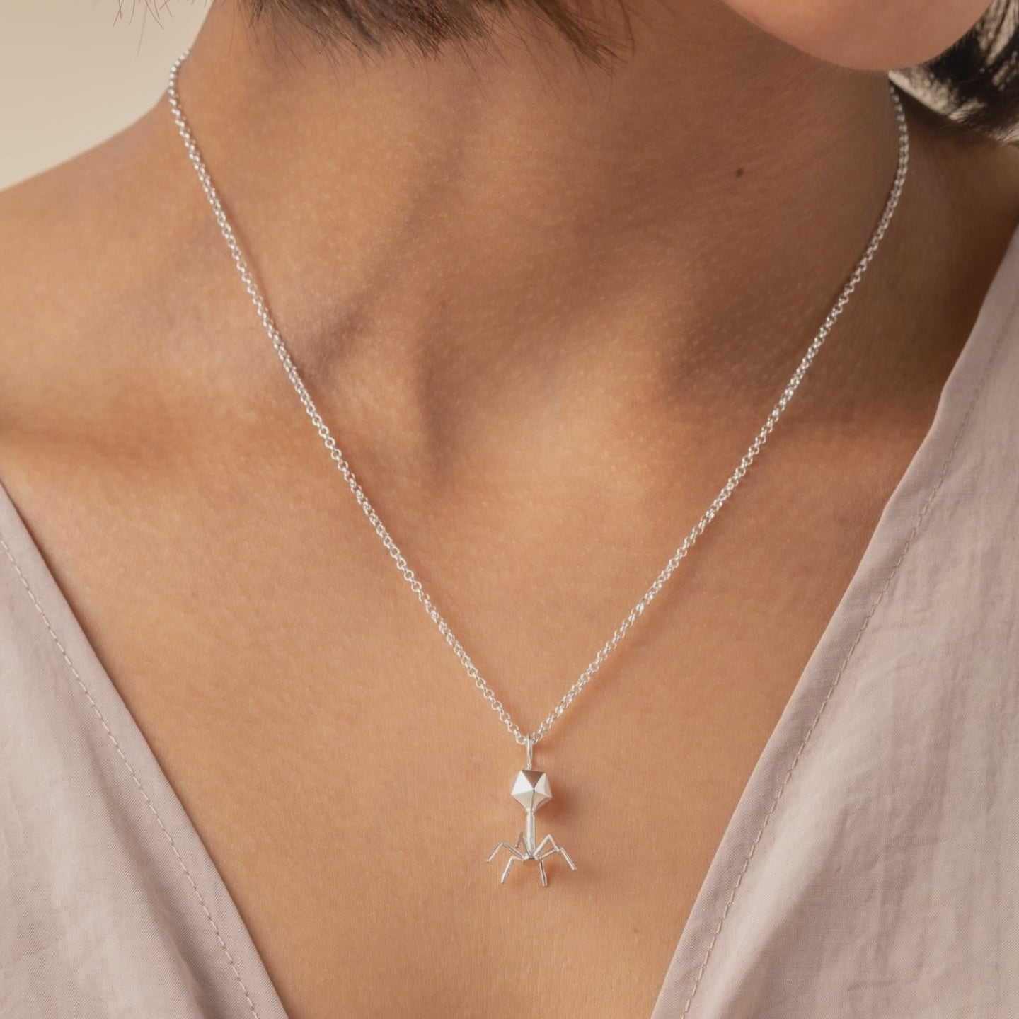 Close-up video of a silver bacteriophage necklace worn on a model, showing the geometric head and delicate leg-like structure as it rests naturally on the skin, with subtle movement and light reflections highlighting the intricate scientific design.