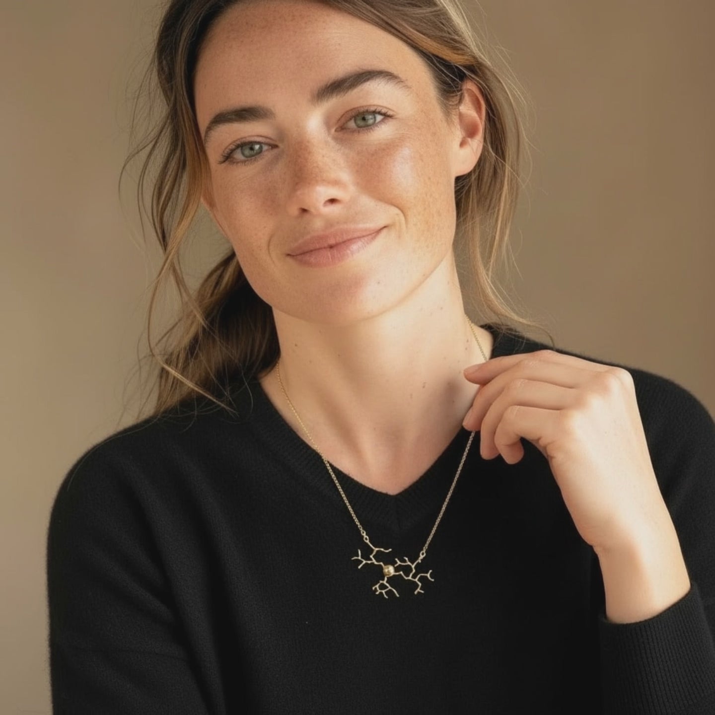 Close-up portrait of a woman wearing a DRG necklace shaped like a branching neuron or molecular structure. She is dressed in a black top and gently holding the chain near her collarbone. The background is warm beige, and soft natural lighting highlights her face and the fine details of the jewelry.