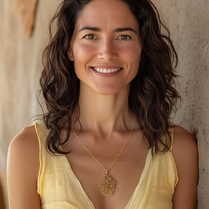 A smiling woman with wavy dark hair wears the gold vermeil Purkinje Cell Necklace. She is dressed in a soft yellow outfit, and the intricate design of the necklace rests just below her collarbone. Her warm expression and natural lighting highlight the elegance of the jewelry.
