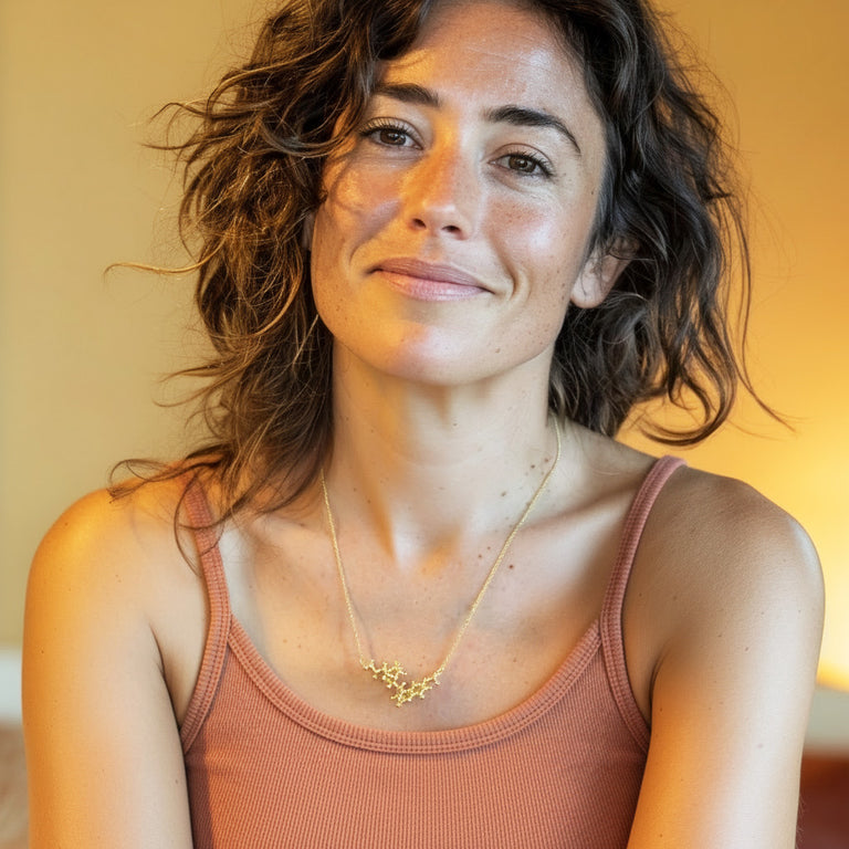 Portrait of a smiling woman wearing a rust-colored tank top and a vermeil gold quinine molecule necklace, shown in warm natural light.