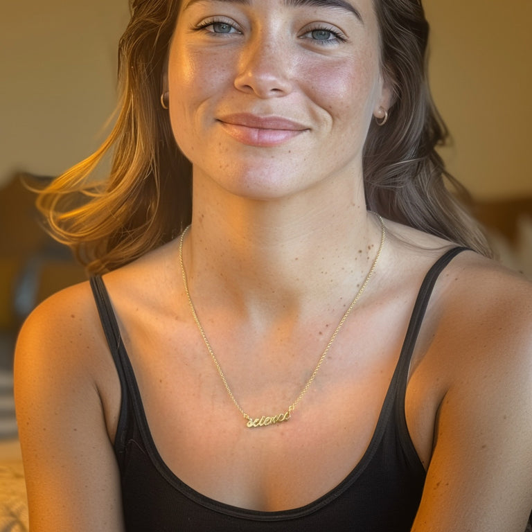 Portrait of a smiling woman wearing the gold vermeil ‘science’ necklace, seated indoors with natural warm lighting.