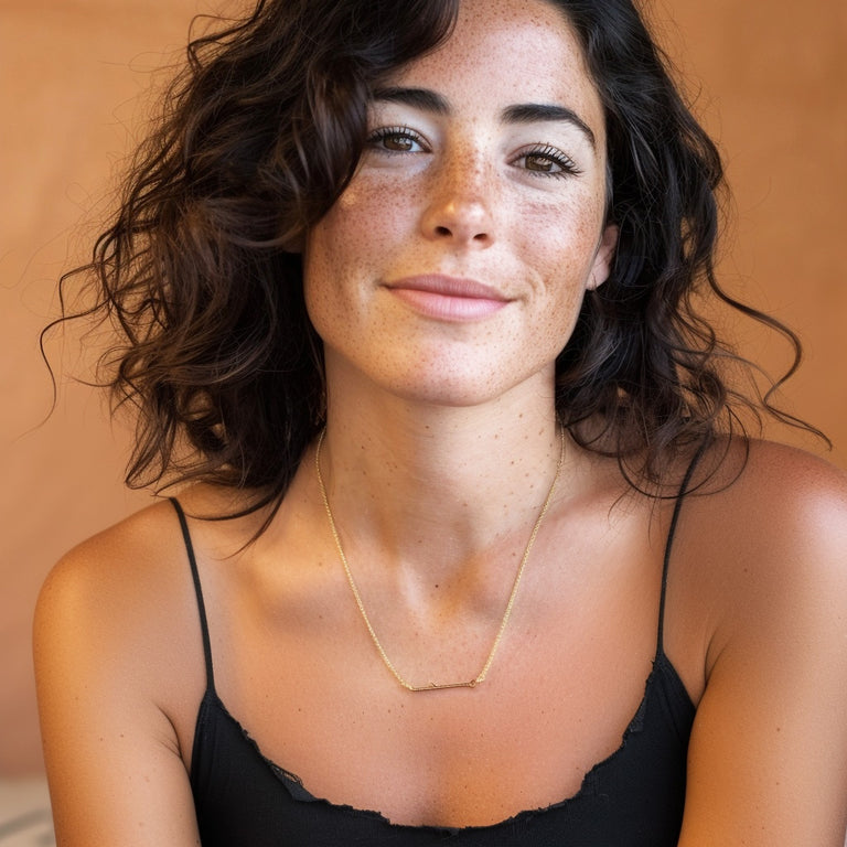 Smiling woman wearing a gold vermeil shRNA necklace, shown from the shoulders up against a warm beige background, highlighting the elegant and minimal pendant on her collarbone.