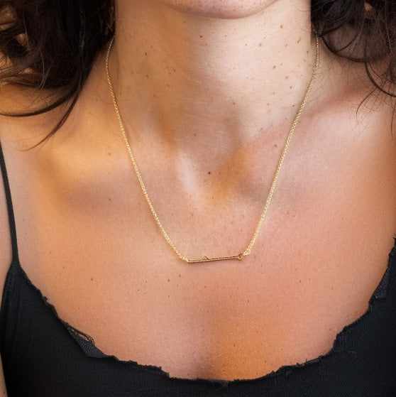 Gold vermeil shRNA necklace worn by a woman, the delicate horizontal pendant resting along her collarbone against a dark V-neck top.