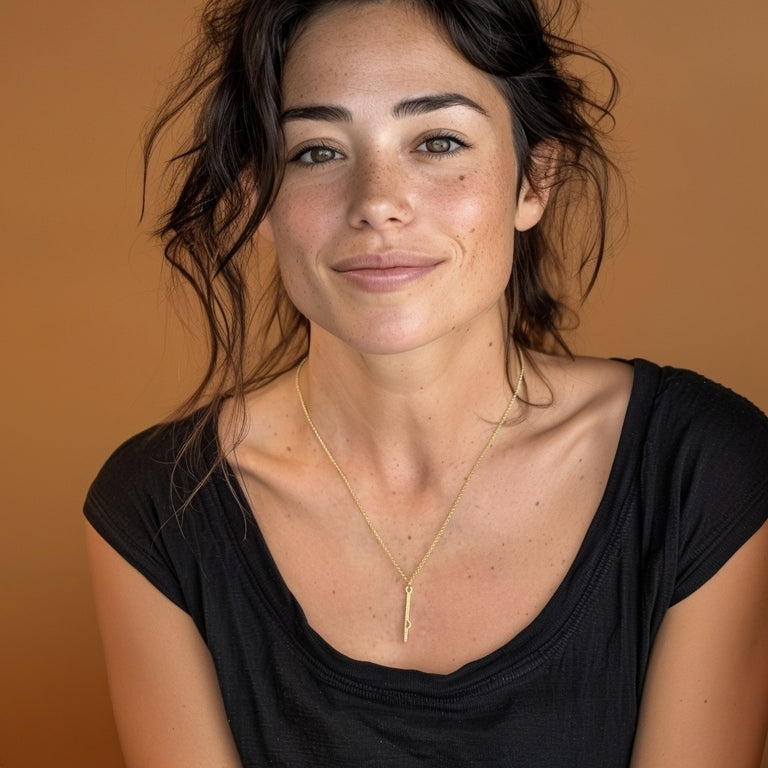 Smiling woman wearing the vertical shRNA gold vermeil necklace against a warm brown background, highlighting the pendant’s elegant and minimalist design.