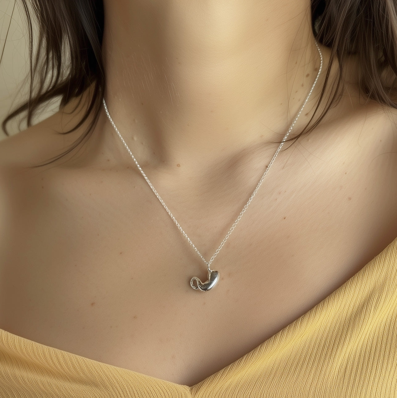 A woman wearing the stomach-shaped necklace. The pendant rests just above her collarbone, suspended on a fine silver chain. She wears a light yellow top, and the image is softly lit.