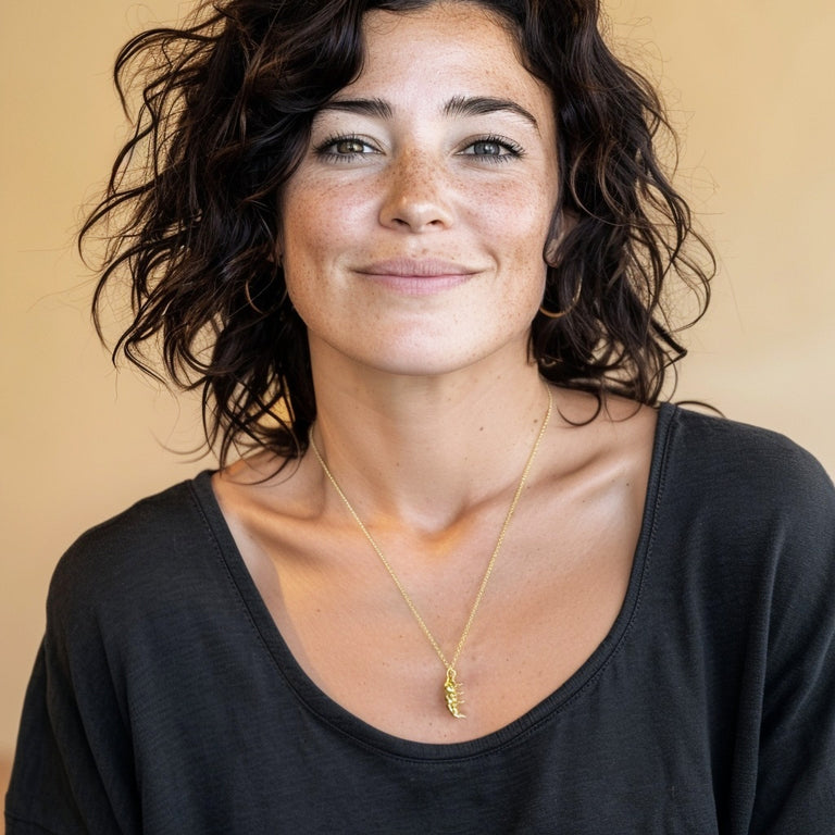Smiling woman with short curly hair wearing the gold vermeil tardigrade necklace, pendant visible against a dark top in a softly lit portrait.