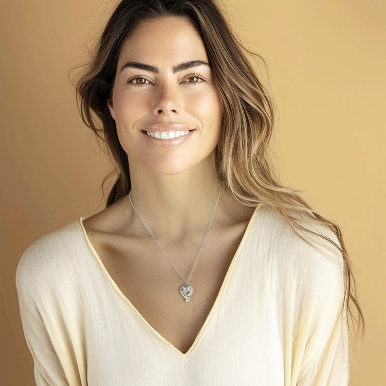 A smiling woman with long, wavy brown hair wearing the sterling silver twin fetus necklace. She is dressed in a soft cream-colored top, and the pendant is prominently displayed against her skin.